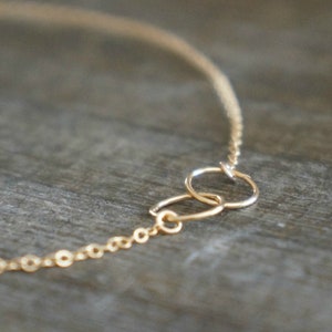 May include: Delicate gold-colored necklace featuring a chain with small circular links. The necklace has two interlocking circles as a focal point. The jewelry is set against a neutral, textured background.