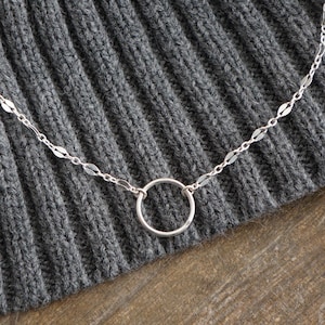 May include: A delicate silver necklace with a small open circle pendant. The chain features small, elongated links and is set against a textured gray background, likely a sweater. The necklace is simple and elegant.