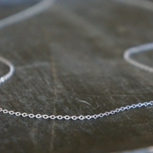 May include: A delicate silver chain necklace with a simple, minimalist design. The chain is composed of small, evenly spaced circular links. The necklace is displayed against a dark, textured background.