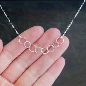 May include: A silver-toned necklace featuring a delicate chain and a central focal point composed of seven interconnected circular rings arranged in a gentle, curved arc. The metal links are thin and polished, creating a simple, continuous design.