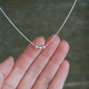 May include: A delicate silver necklace featuring three small, round beads. The beads are strung on a fine silver chain. The necklace is displayed against a soft, neutral background.
