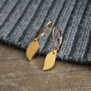 May include: A pair of gold-colored leaf-shaped earrings. The earrings have a lever-back closure and are displayed on a gray knitted fabric. The earrings are a simple, elegant design.