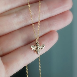 May include: A delicate gold-colored necklace featuring a small bee pendant. The bee pendant has detailed wings and a crown. The necklace chain is thin and appears to be approximately 18 inches long.