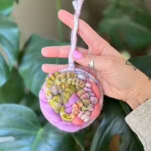 May include: A hand holds a colorful yarn ball ornament with a pink, green, and yellow color scheme. The ornament is made of woven yarn and has a loop for hanging.
