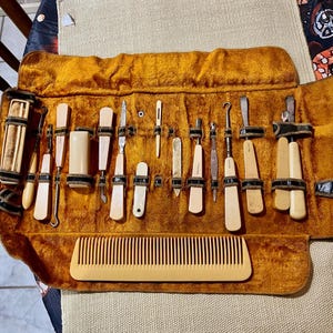 Pode incluir: Um antigo conjunto de manicure com várias ferramentas de cor marfim e um pente, apresentado numa caixa de veludo mostarda. O conjunto inclui limas de unhas, empurradores de cutículas e outros instrumentos de higiene, todos presos com correias pretas.
