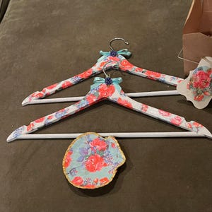 May include: Two white wooden clothes hangers with floral patterns in red, blue, and green. Each hanger has a silver hook, a light blue bow, and a small blue decorative element. A small, decorative, shell-shaped dish with a floral pattern is also present.