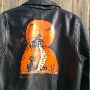 May include: Black leather jacket with an orange and black Halloween-themed design painted on the back. The design features a haunted house, a black cat, a pumpkin, and the text "Kustom Artistry".