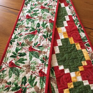 May include: A quilted table runner featuring a white fabric with red cardinals, green pine branches, and brown pinecones on one side, and a patchwork design of green, red, and yellow squares on the other, finished with a red fabric border.
