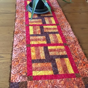 May include: A quilted table runner with a geometric pattern in shades of orange, yellow, brown, and pink. The runner has a brown border and is decorated with a small potted plant.