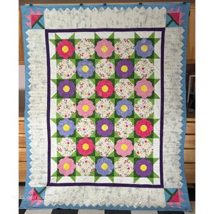 May include: A rectangular quilt featuring a grid of alternating floral-patterned squares and solid-colored flower shapes in pink, purple, and yellow. The quilt has a dark purple inner border and a light blue outer border with pink flower corners.