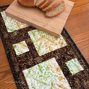 May include: A rectangular quilted table runner featuring a batik pattern with dark brown and white sections. The white fabric displays a green and yellow botanical print. The text The Quilted Bungalow is printed in the bottom left corner.