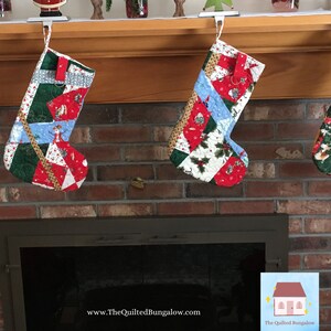 May include: Two quilted Christmas stockings hang from a wooden mantel. The stockings feature patchwork designs with red, green, and blue fabrics. One stocking has a snowman, and the other has holly and berries. The website address www.TheQuiltedBungalow.com is visible.