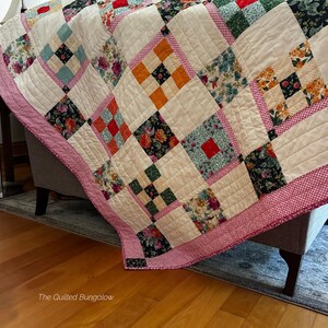 May include: A patchwork quilt featuring a variety of floral and solid-colored fabric squares in shades of orange, red, green, and black, separated by cream-colored quilted blocks and pink patterned borders. The text The Quilted Bungalow is visible.
