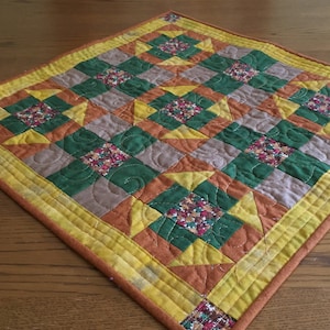 May include: A small quilted table runner with a geometric pattern in shades of green, orange, yellow, and brown. The runner is made of fabric squares sewn together to create a patchwork design. The text "The Quilted Bungalow" is visible in the bottom right corner of the image.