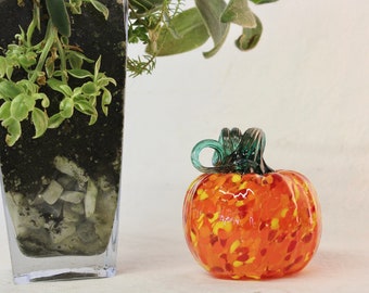 May include: A glass pumpkin with a green stem. The pumpkin is orange with yellow and red speckles. The pumpkin is sitting on a white surface.