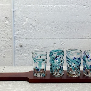 May include: A wooden serving paddle holding four shot glasses. The glasses feature a blue and teal swirl design. The paddle is dark brown and has a hole in the handle. The glasses are for serving drinks.