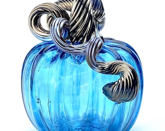 May include: A decorative glass pumpkin in a vibrant blue hue. The pumpkin features a textured surface and is topped with a silver-colored, swirled stem. This handcrafted piece is a unique fall or autumn-themed home decor item.