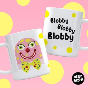 May include: Two white ceramic mugs with quirky designs. One mug features a pink blob character with a yellow bow tie and a smiling face. The other mug has the word "Blobby" repeated three times. Yellow polka dots are scattered on the mugs.