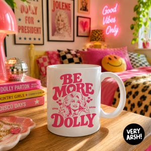 May include: A white ceramic mug featuring a pink graphic illustration of Dolly Parton with the text "BE MORE DOLLY" and the years "1946" and "2026" printed on the front. The design includes small heart accents surrounding the portrait.