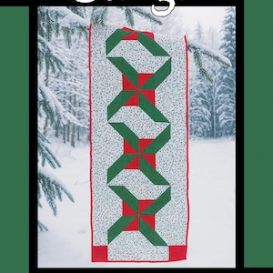 May include: A Christmas-themed quilt pattern with the word "Jingle" at the top. The quilt features a repeating pattern of green and red shapes on a white background with red borders. The quilt is hanging on a snowy tree branch.