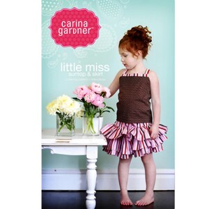 May include: A girl wearing a brown and white polka dot tank top and a pink and white striped ruffle skirt. The girl is holding a bouquet of pink and white flowers. The image also includes a white table with two clear glass vases filled with flowers. The text "carina gardner" and "little miss sun top & skirt" is visible in the image.