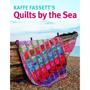 May include: A book cover titled Kaffe Fassett's Quilts by the Sea featuring a vibrant, multi-colored patchwork quilt with floral and geometric patterns draped over a weathered, pink and brown wooden boat on a pebble beach by the ocean.