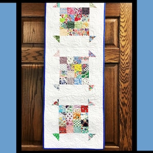 May include: A rectangular table runner featuring three square patchwork blocks made of multi-colored floral and geometric fabric scraps set against a white quilted background with swirling patterns and a solid blue border. Text reads Nana's Aprons.