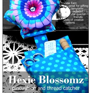 May include: A blue polka-dot pincushion and thread catcher set with a star-shaped pincushion featuring a floral design in shades of blue, purple, and pink. The set includes scissors and a wooden spool. Text reads "Hexie Blossomz".
