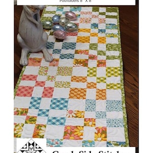 May include: A Charming Four Patch Twist quilt runner measuring 16 inches by 48 inches, with a trivet at 16 inches by 16 inches and potholders at 8 inches by 8 inches. The quilt features a multi-colored patchwork pattern with floral and gingham prints.