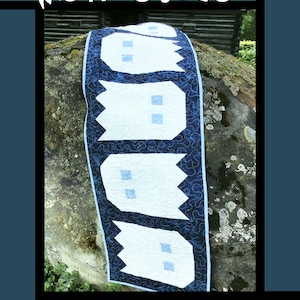 May include: A blue and white quilt with a pattern of ghosts. The quilt is on a large rock. The text "Ghosties" is at the top of the image. The text "Tricia Lynn Maloney Orphan Quilt Designs For Villa Rosa Designs" is at the bottom of the image.