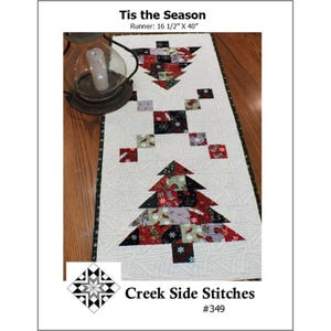 May include: A rectangular table runner titled Tis the Season measuring 16 1/2 inches by 40 inches. The white quilted fabric features two patchwork Christmas tree designs made of red, black, and green patterned squares with a dark border.