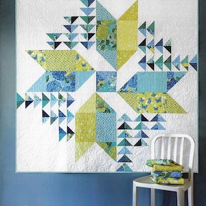 May include: A white quilt with a starburst design in shades of blue, green, and yellow hangs on a blue wall. The quilt is labeled "Wind Drifter" and is a quilt pattern by Robin Pickens. A white chair holds folded fabric.