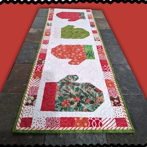May include: A quilted table runner with a white background and colorful mitten designs. The mittens are red, green, and patterned with snowflakes and floral prints. The border is red and white with a green edge. Text at the top reads "COLOR MITTENS".