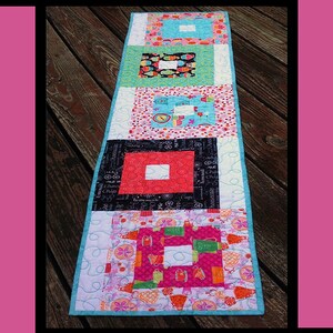 May include: A colorful table runner with a cupcake party theme. The runner is made of fabric squares with different patterns and colors, including pink, blue, green, black, and white. The runner is quilted and has a white background. The text "Cupcake Party" is printed in black at the top of the image. The text "Tricia Lynn Maloney Orphan Quilt Designs For Villa Rosa Designs" is printed at the bottom of the image.