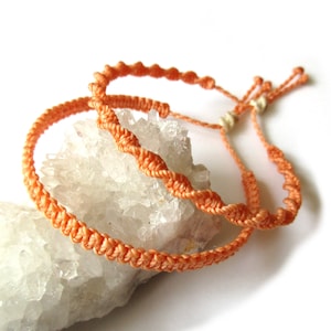 Könnte beinhalten: Zwei geflochtene Armbänder in Pfirsichfarbe mit einem geknoteten Verschluss. Die Armbänder werden auf einem weißen Kristall präsentiert.