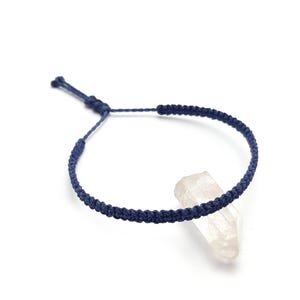 Peut inclure: Un bracelet macramé bleu marine avec une fermeture coulissante réglable. Le bracelet est présenté sur fond blanc, à côté d'un cristal de quartz clair. Le bracelet est de conception simple et minimaliste.