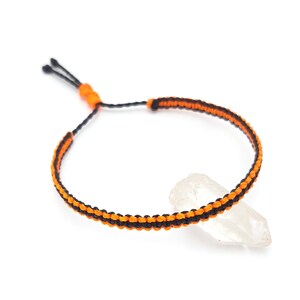 Peut inclure: Un bracelet macramé noir et orange avec une fermeture coulissante réglable. Le bracelet en cordon tressé est posé sur un cristal clair. Le design présente une base noire avec des détails orange. Le bracelet mesure environ 15-25 cm de circonférence.
