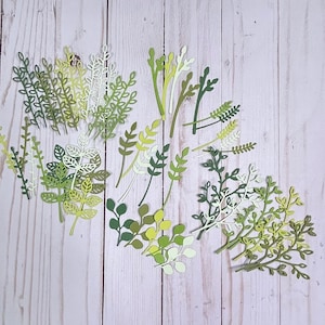 May include: Assortment of green, yellow, and white paper leaves and branches. The leaves are cut out in various shapes and sizes, and some have a delicate, lacy design. The leaves are arranged on a light wood background.