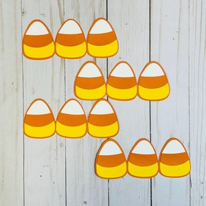 May include: A set of ten paper candy corn cutouts in orange, yellow, and white. The cutouts are arranged in a staggered pattern on a light wood background.