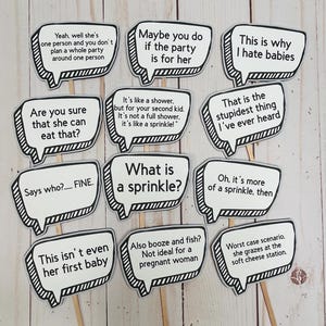 May include: A collection of speech bubble-shaped party decorations with various humorous phrases printed in black text. Each speech bubble is outlined in black and mounted on a wooden stick. The phrases include funny sayings about babies and parties.
