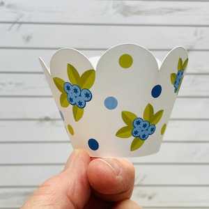 May include: White cupcake liner with a scalloped edge and a blue and green floral pattern. The pattern features blueberries and leaves.