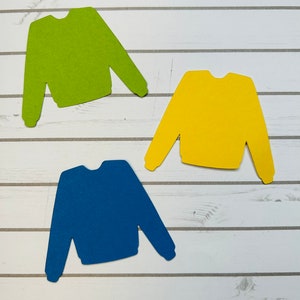 May include: Three paper cutouts of sweaters in different colors: green, yellow, and blue. The sweaters are on a white wood background.