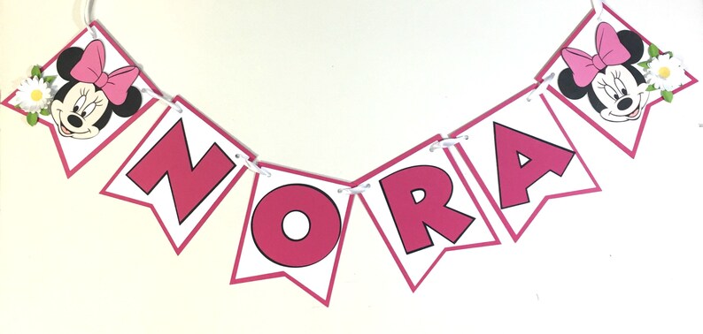 Puede incluir: Una pancarta rosa y blanca con el nombre "Nora" escrito en letras rosas. La pancarta est&aacute; decorada con dos cabezas de dibujos animados de Minnie Mouse con lazos rosas y flores blancas.