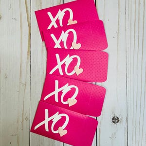 May include: Five pink envelopes with white "XO" and a pink heart on each. The envelopes are stacked on top of each other.