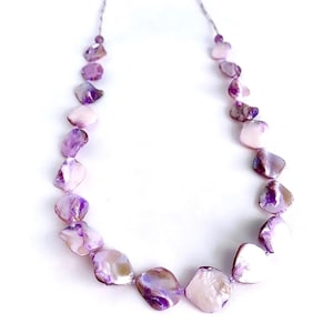 以下が含まれることがあります： 銀色のチェーンに、紫と白の貝殻のネックレス。ネックレスは、不規則な形の貝殻のビーズで作られています。