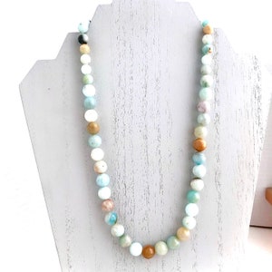 May include: A light blue, green, and brown beaded necklace made of natural stones. The necklace is displayed on a white jewelry stand.