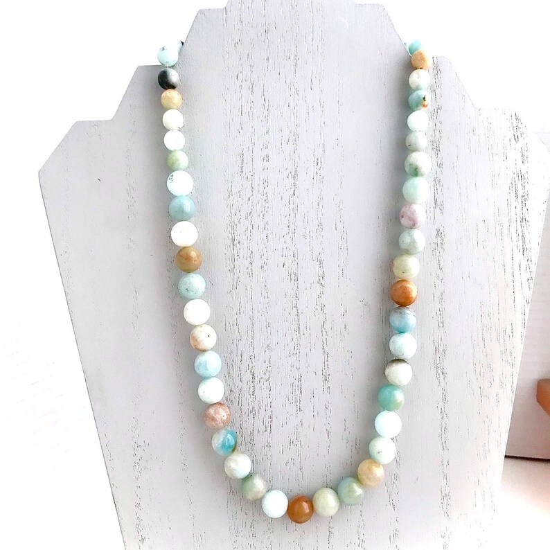 May include: A light blue, green, and brown beaded necklace made of natural stones. The necklace is displayed on a white jewelry stand.