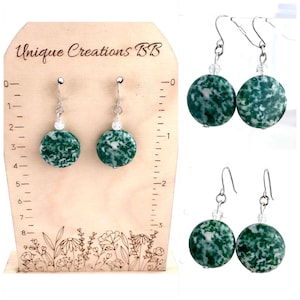 May include: Green and white speckled stone earrings with silver-colored hooks and a small clear bead accent. The earrings are displayed on a wooden stand with the text "Unique Creations BB". Additional earrings are shown in the image.