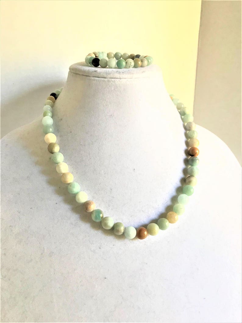 May include: A green, brown, and white beaded necklace and bracelet set. The necklace is on a white mannequin bust. The beads are round and have a natural, polished look.