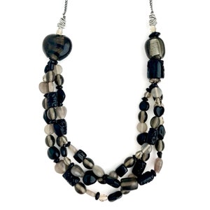 May include: A multi-strand necklace with black and gray beads. The necklace features a heart-shaped bead at the top and a variety of other shapes, including round, oval, and square beads.